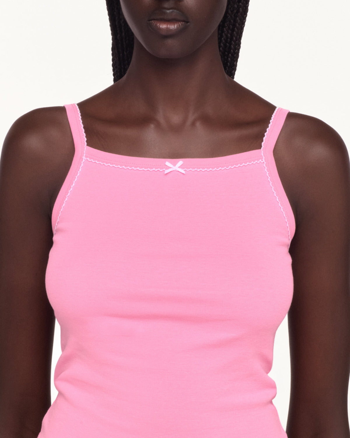 A close-up of a model wearing a light pink ribbed camisole tank top with thin straps, white contrast stitching, and a small white bow at the center of the neckline.
