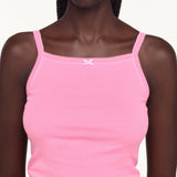 A close-up of a model wearing a light pink ribbed camisole tank top with thin straps, white contrast stitching, and a small white bow at the center of the neckline.
