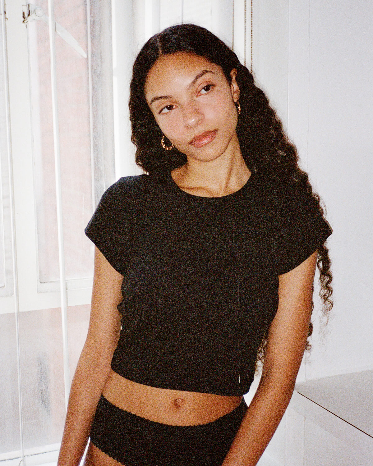 Woman wearing a black crop top and black shorts indoors with white curtains in the background