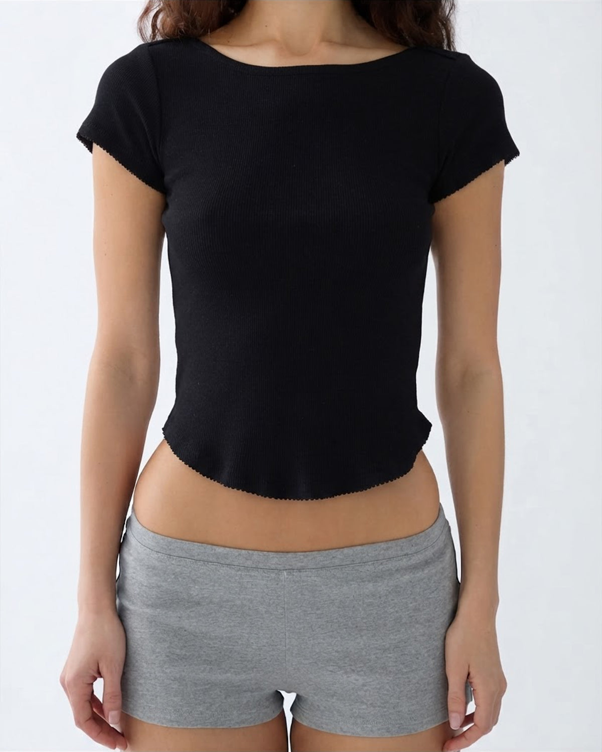A woman wearing a black short-sleeve boat-neck crop top and gray athletic shorts, shown from the neck down against a plain light gray background.