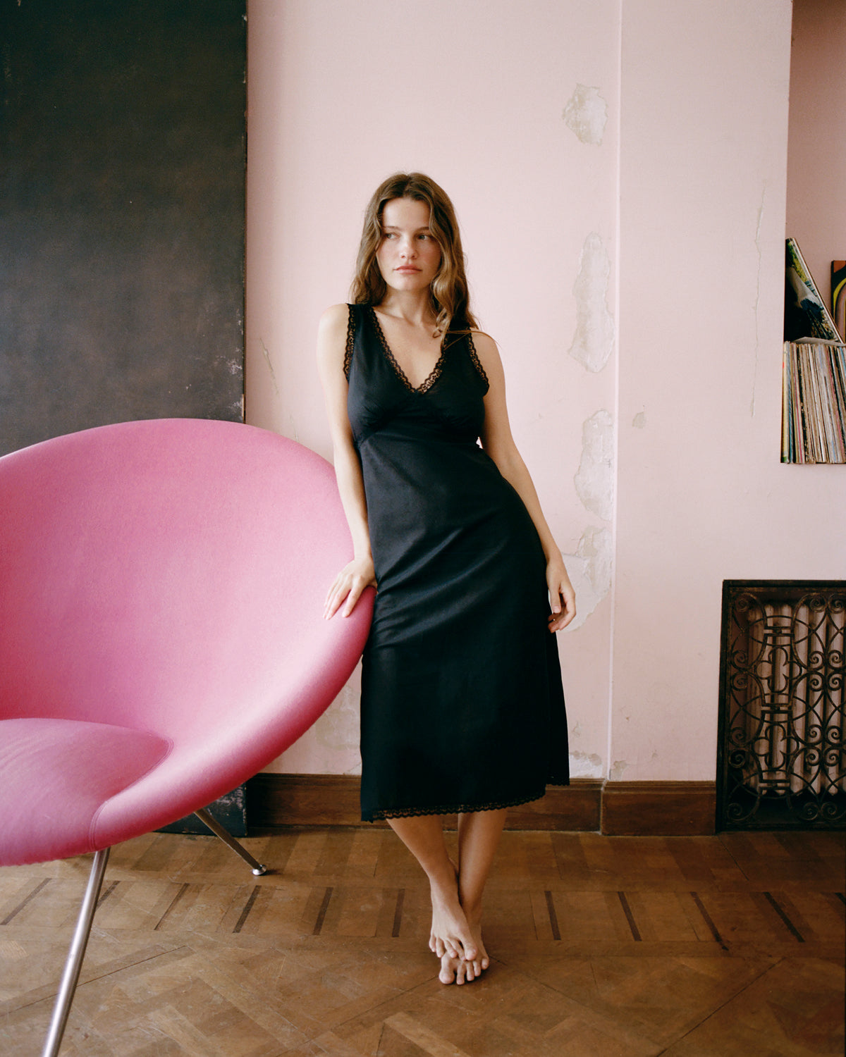 Woman in a black dress standing next to a pink chair in a room with a pink wall.