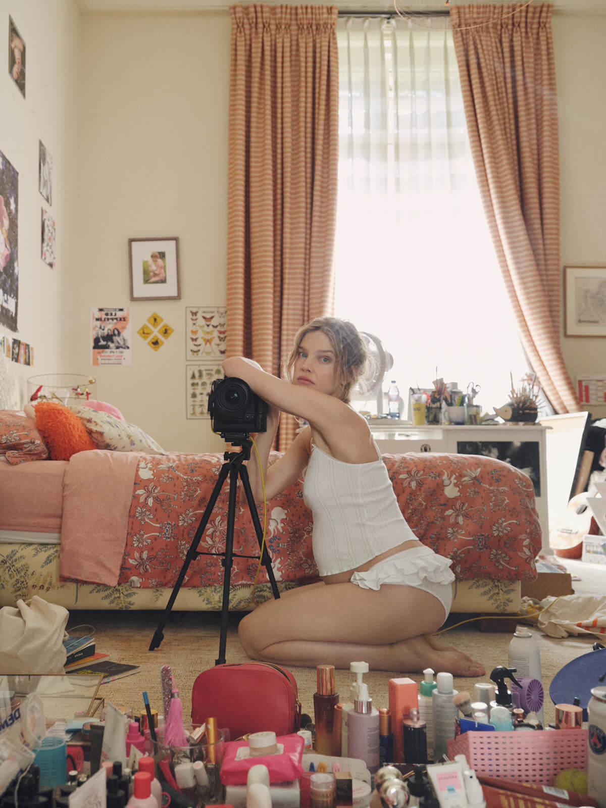 A woman kneels on the floor in a bedroom setting, holding a vintage-style camera on a tripod. She is wearing a white camisole and ruffled shorts.