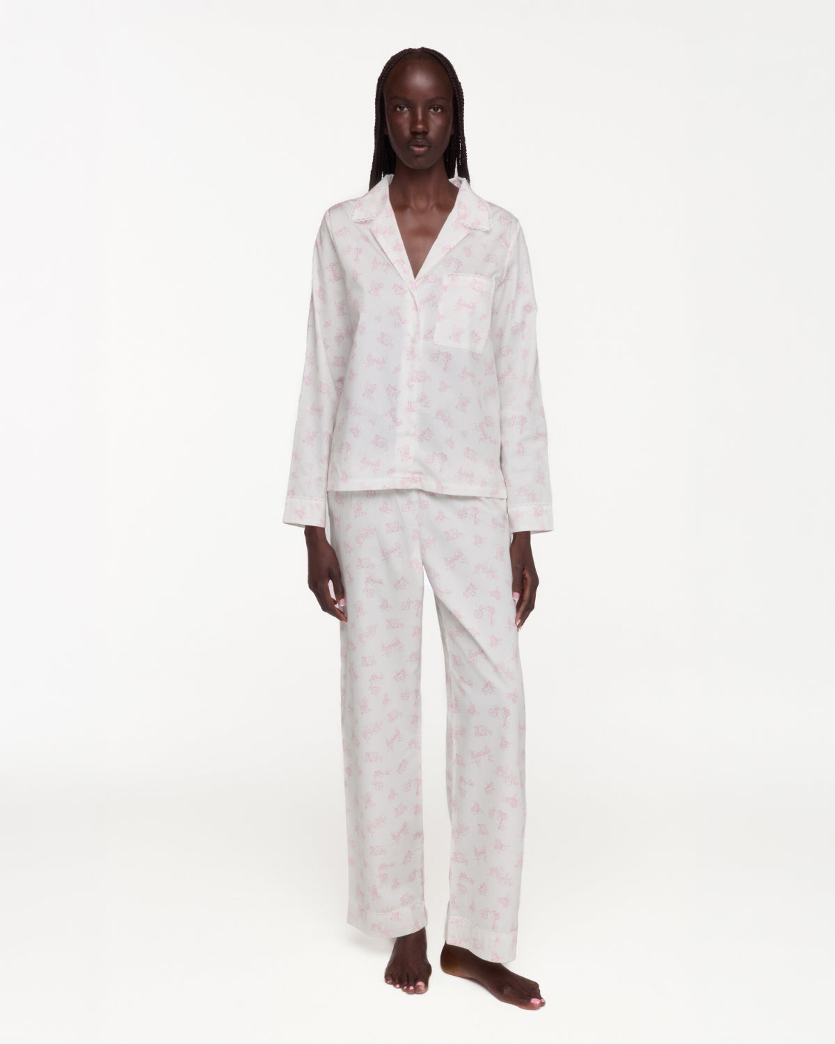 A model stands against a plain white studio background wearing a matching pajama set with a light pink pattern on a white base. The set includes a long-sleeve button-front top with a chest pocket and matching wide-leg pants.