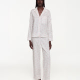 A model stands against a plain white studio background wearing a matching pajama set with a light pink pattern on a white base. The set includes a long-sleeve button-front top with a chest pocket and matching wide-leg pants.