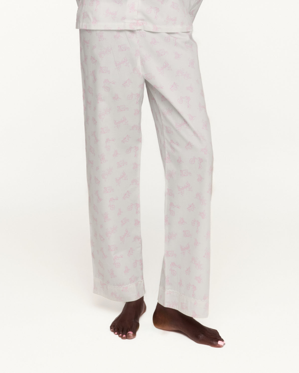A person wearing white pajama pants with a small pink repeating pattern, shown from the waist down against a plain white studio background.