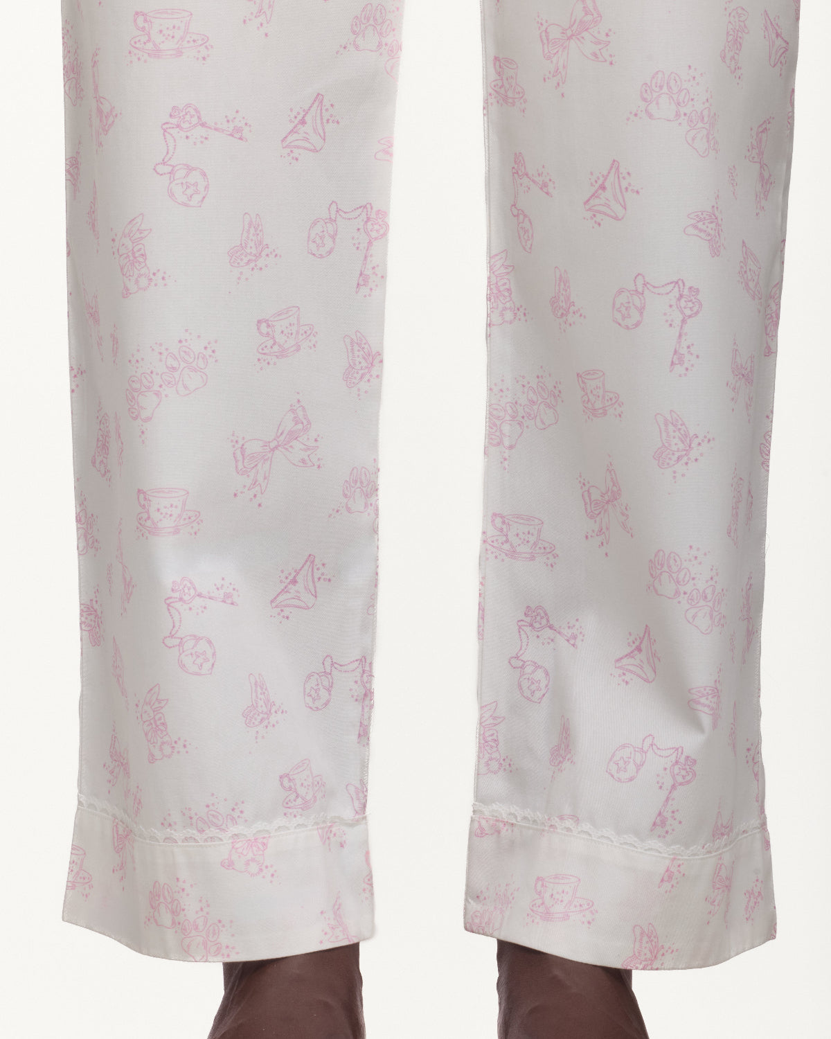 Close-up of a pair of white trousers with a delicate pink whimsical pattern and a lace trimmed hem, shown on a model's legs.
