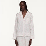 A model wearing a light pink and white patterned pajama set, consisting of a long-sleeve button-up top and matching pants, against a plain white studio background.