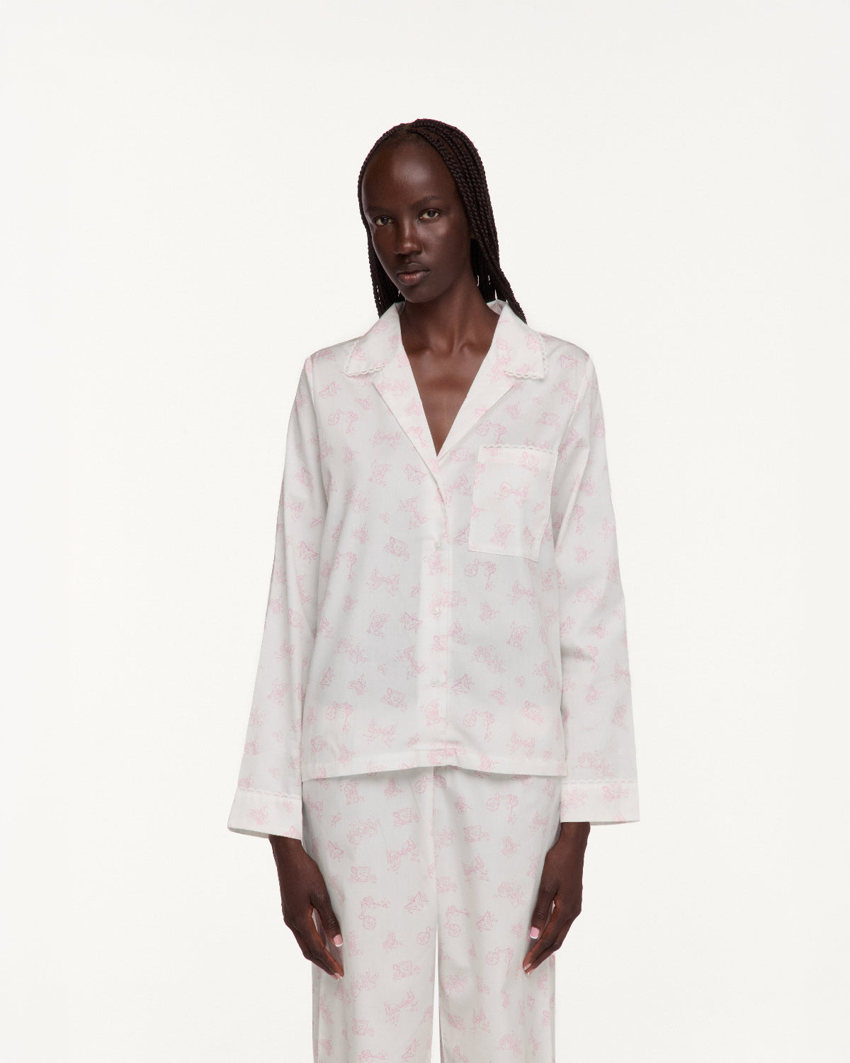 A model wearing a light pink and white patterned pajama set, consisting of a long-sleeve button-up top and matching pants, against a plain white studio background.
