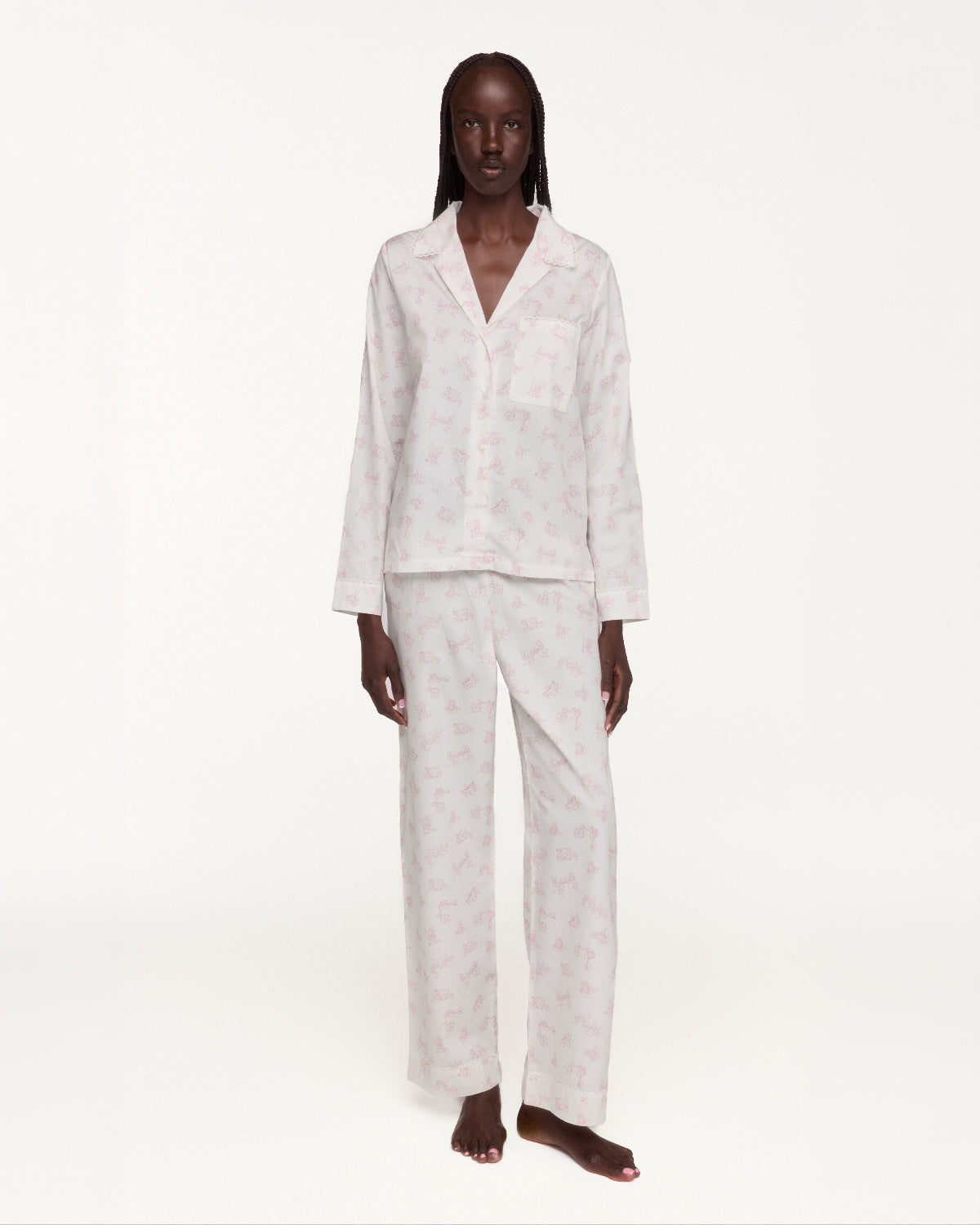 A model stands against a plain white studio background wearing a matching pajama set consisting of a long-sleeve button-front top with a chest pocket and coordinating wide-leg pants. The pajamas are white with a subtle repeating floral print.