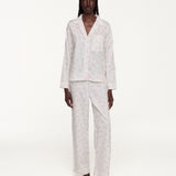 A model stands against a plain white studio background wearing a matching pajama set consisting of a long-sleeve button-front top with a chest pocket and coordinating wide-leg pants. The pajamas are white with a subtle repeating floral print.