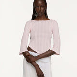 A model wearing a light pink three-quarter sleeve crop top and a white skirt, photographed against a solid off-white studio background.