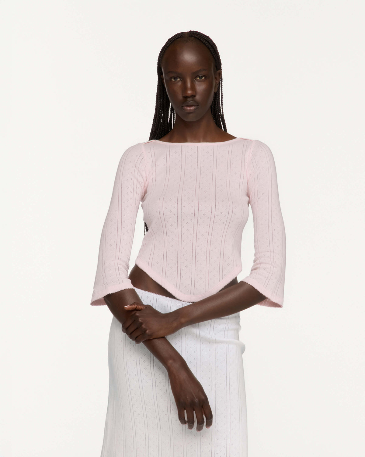 A model wearing a light pink three-quarter sleeve crop top and a white skirt, photographed against a solid off-white studio background.