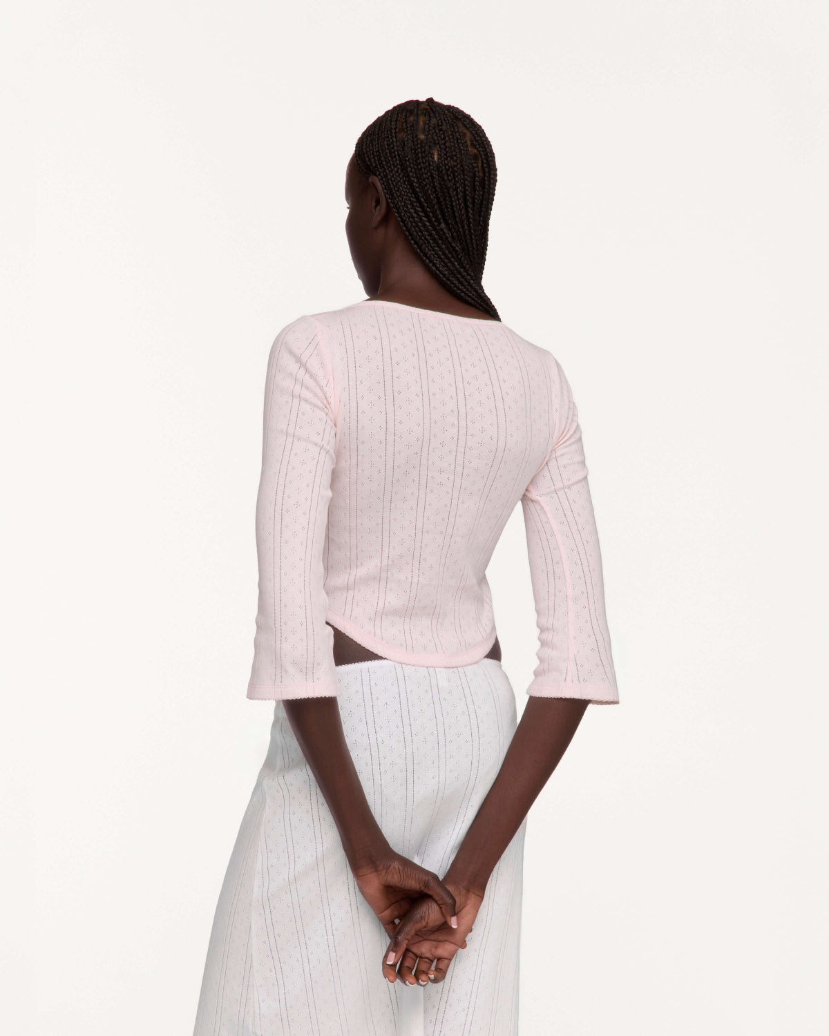 A woman shown from the back wearing a light pink three-quarter sleeve top and a white pleated skirt, set against a plain off-white studio background.