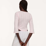 A woman shown from the back wearing a light pink three-quarter sleeve top and a white pleated skirt, set against a plain off-white studio background.