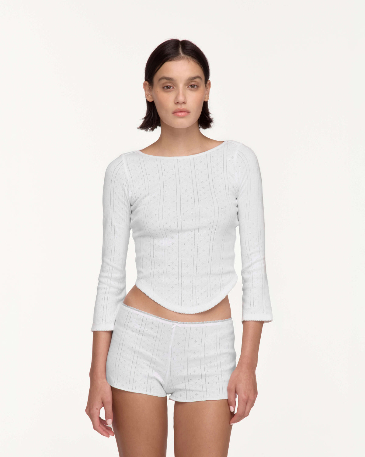 A model wearing a white three-quarter sleeve top and matching white shorts against a plain off-white studio background.