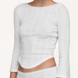 A woman wearing a white three-quarter sleeve top with a curved hemline, paired with matching white bottoms against a light off-white background.