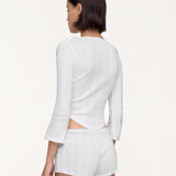 A model shown from the back wearing a white long-sleeve crop top and matching white shorts against a plain off-white studio background.
