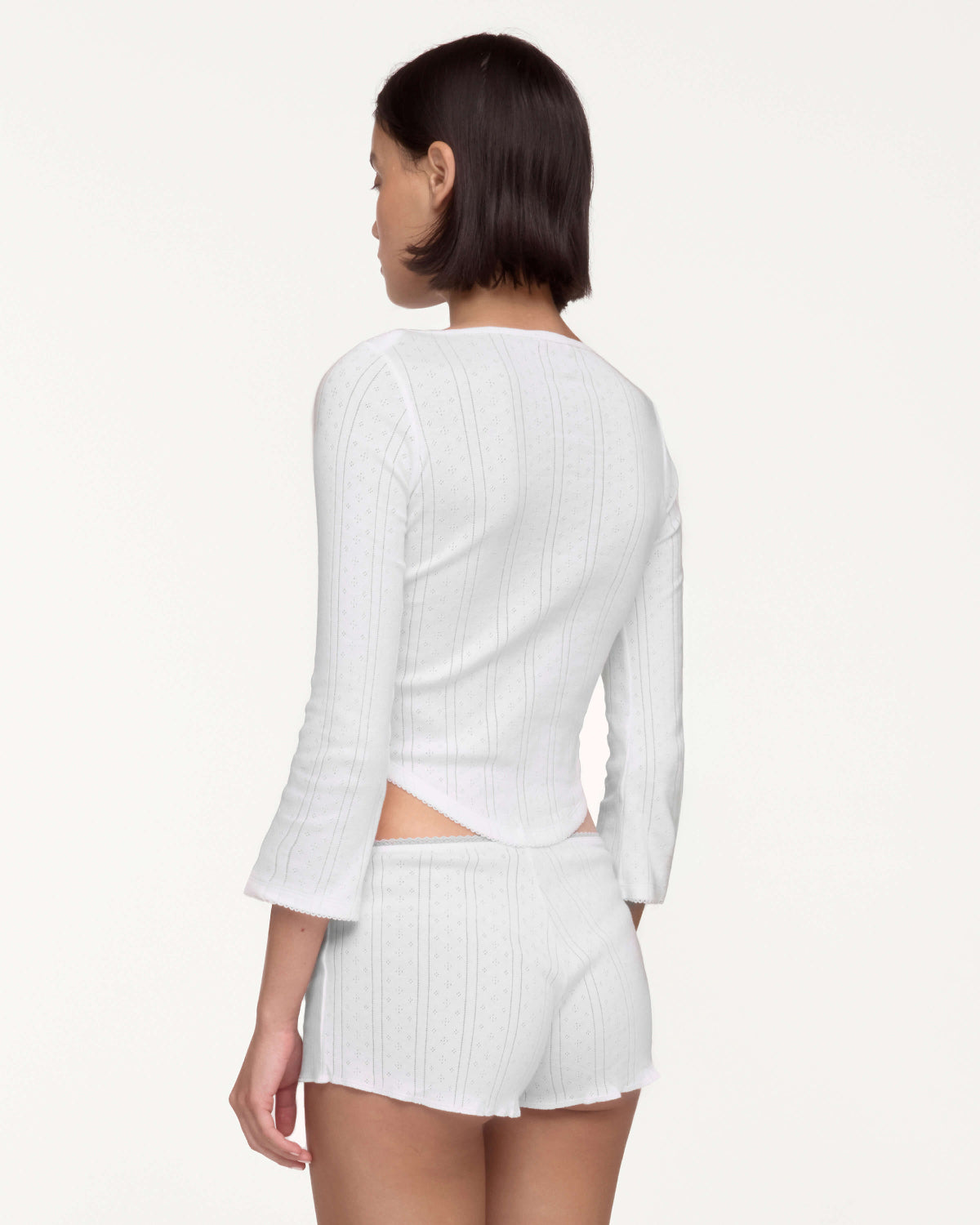 A model shown from the back wearing a white long-sleeve crop top and matching white shorts against a plain off-white studio background.