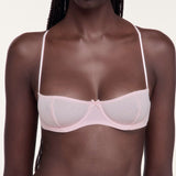 A close-up of a model wearing a light pink underwire bra with thin straps and a center bow detail, set against a plain light background.