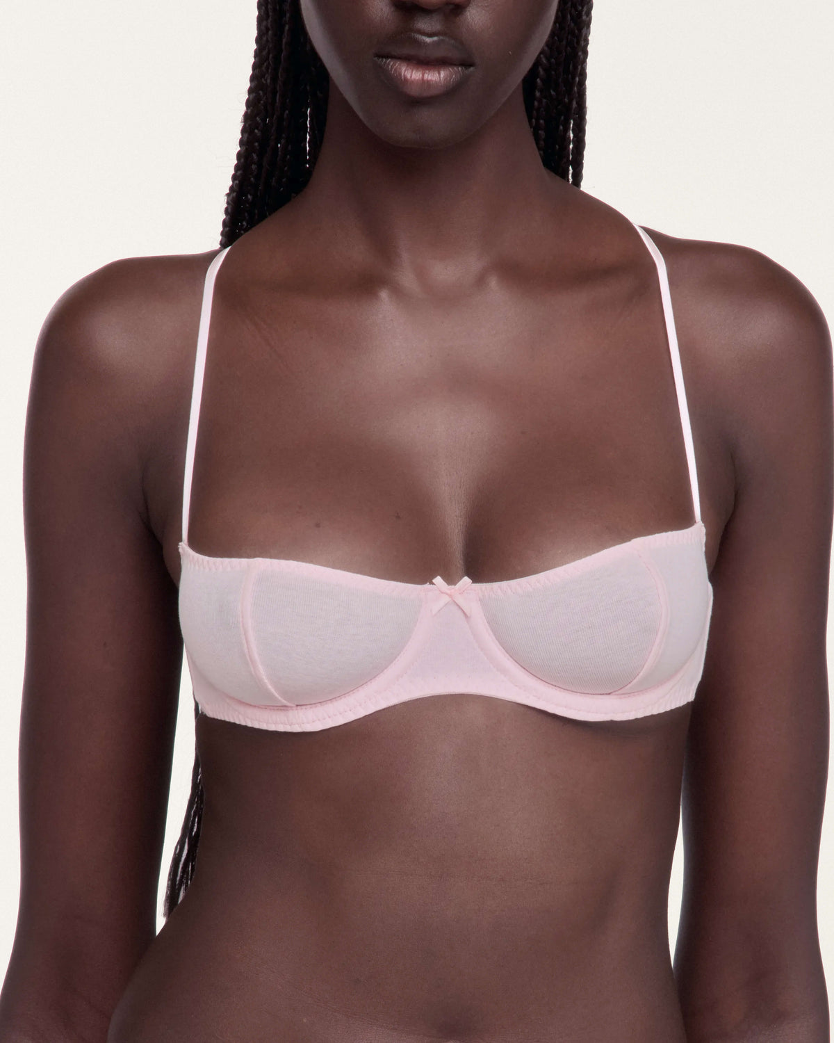 A close-up of a model wearing a light pink underwire bra with thin straps and a center bow detail, set against a plain light background.