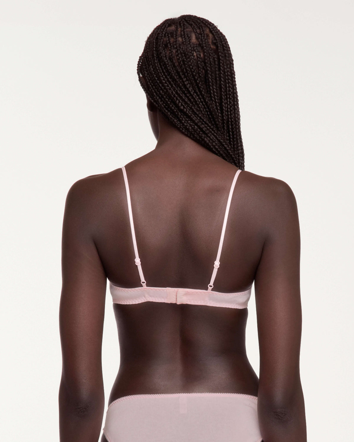 A woman viewed from the back wearing a pale pink bralette with thin adjustable straps and a matching light pink bottom, against a plain light background.