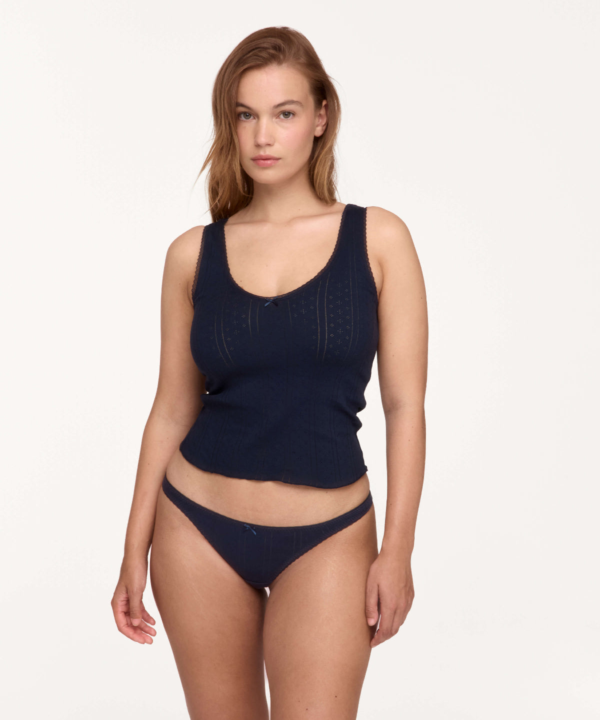 A woman modeling a dark navy blue ribbed knit two-piece set consisting of a scoop-neck tank top and matching low-rise briefs, shot against a solid off-white studio background.