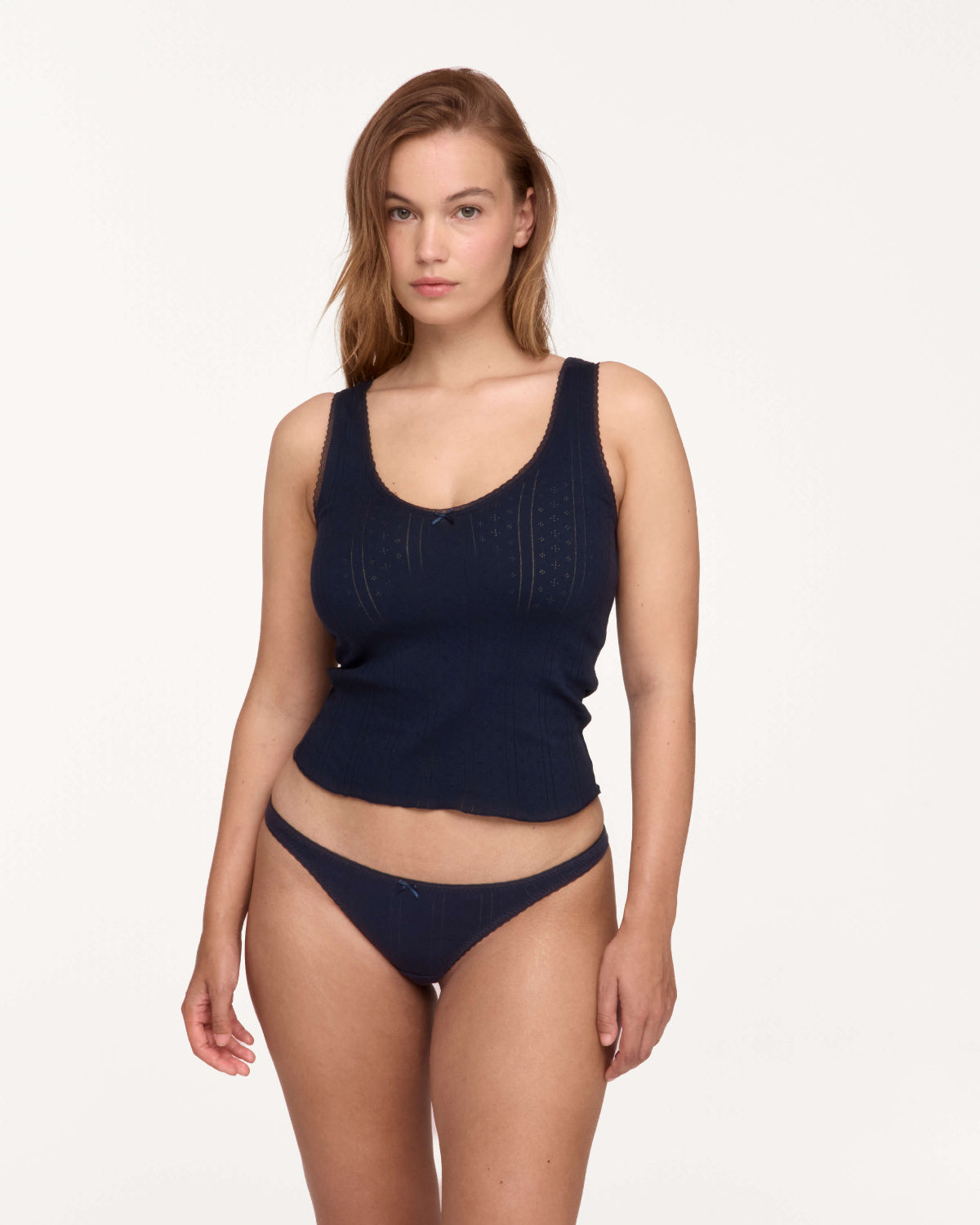 A woman modeling a dark navy blue ribbed knit two-piece set consisting of a scoop-neck tank top and matching low-rise briefs, shot against a solid off-white studio background.