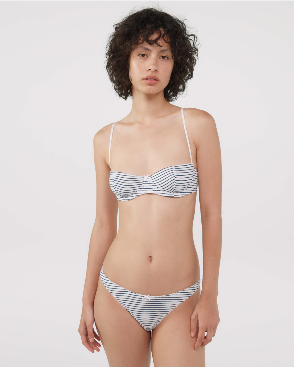 A model wearing a bra and thong with thin white straps and a blue and white horizontal stripe pattern, photographed against a solid light gray background.