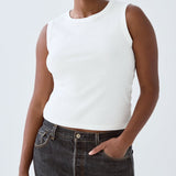 Person wearing a white sleeveless top and dark jeans on a plain background