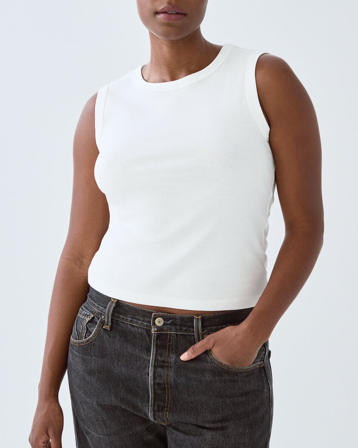 Person wearing a white sleeveless top and dark jeans on a plain background