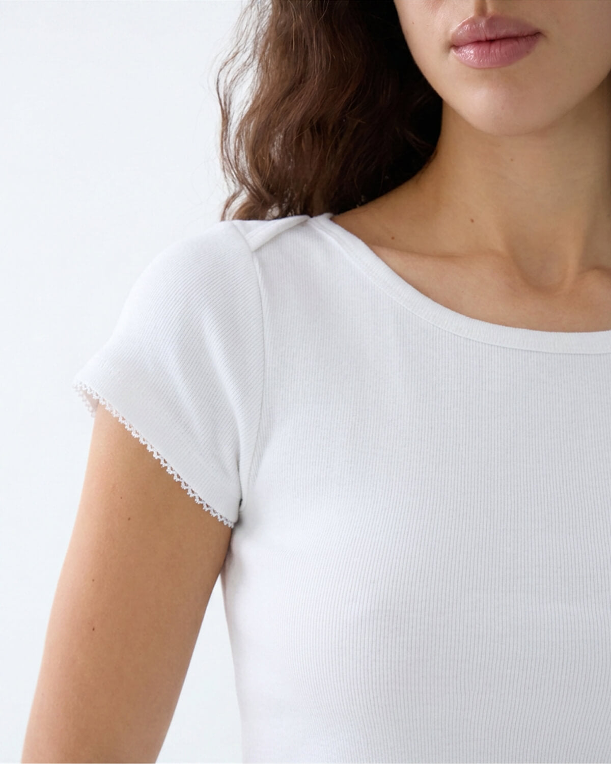Close-up of a woman wearing a white short-sleeve ribbed top with delicate scalloped lace trim at the sleeve hems, set against a plain white background.