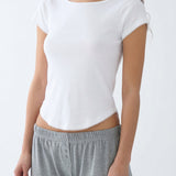 A woman wearing a white short-sleeve scoop-neck top with delicate scalloped edges and grey lounge pants, shown from the neck down against a plain white background.